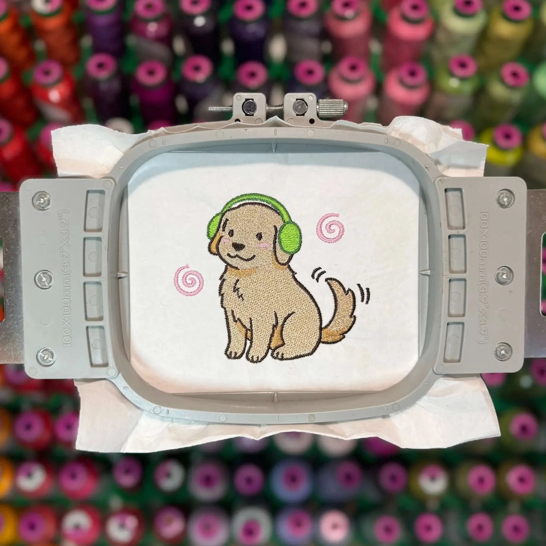 Embroidery hoop with a cartoon dog wearing headphones on a white fabric background, against a colorful spool of thread backdrop.